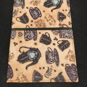 May include: A brown fabric journal cover with a coffee-themed pattern. The design includes coffee pots, mugs, and beans, along with the words "Coffee" and other coffee-related phrases. A black elastic band secures the cover.
