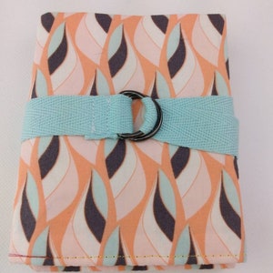 Peut inclure: Une petite pochette rectangulaire en tissu avec une sangle bleu clair et un anneau métallique. La pochette est en tissu de couleur pêche avec un motif de feuilles blanches et turquoise.
