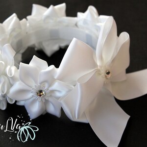 May include: White satin headband with a large bow and several smaller flower-shaped embellishments. Each flower has a small rhinestone in the center.