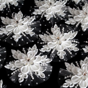 Puede incluir: Primer plano de múltiples adornos florales de tela blanca con un brillante central y detalles de perlas. Las flores están colocadas sobre un respaldo de red transparente con lunares, creando un diseño delicado y elegante. Ideal para proyectos nupciales o de manualidades.