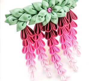 May include: A handcrafted hair accessory featuring a floral design with cascading ribbons. The piece showcases a gradient of pink hues, transitioning from deep magenta to pale pink, complemented by green leaves and a central pink gem.