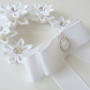 May include: White fabric flower wreath with a white satin ribbon bow and a pearl and rhinestone embellishment.