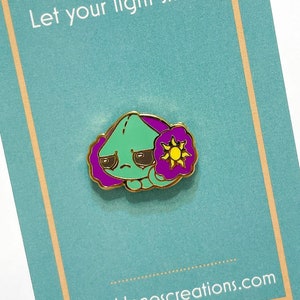 May include: A green enamel pin with a sad face and purple flower-like accents, and a yellow sun. The pin is on a teal card with the text "Let your light shine!" and the website kathlenescreations.com.