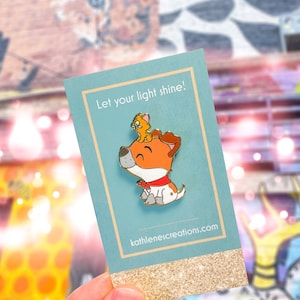 Pode incluir: Pin de esmalte com um cão de desenho animado com um pequeno animal nas costas, sobre um fundo verde-azulado. O pin tem uma coleira vermelha e o texto "Let your light shine!" está impresso no cartão. O site kathlenescreations.com também é visível.