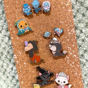 May include: A collection of enamel pins featuring various cartoon characters. The pins include a blue character with a top hat, a cat wearing a witch hat on a broom, and a cat in a dress. The pins are set on a corkboard.