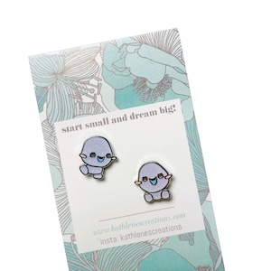 May include: A pair of purple and blue enamel pin earrings featuring a cartoon character with a smiling face. The earrings are on a white card with the text "start small and dream big!" and the website and Instagram handle for the seller.