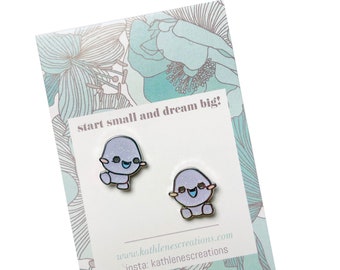 May include: A pair of purple and blue enamel pin earrings featuring a cartoon character with a smiling face. The earrings are on a white card with the text "start small and dream big!" and the website and Instagram handle for the seller.