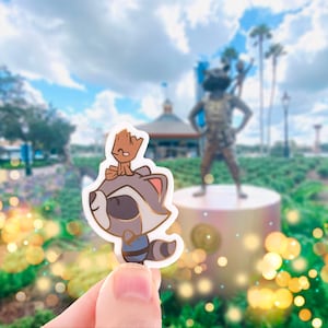 May include: A sticker featuring Rocket Raccoon and Baby Groot from Guardians of the Galaxy. Rocket is gray, white, and brown, with a blue outfit. Baby Groot is brown and sits on Rocket's head. The background is a blurred outdoor scene.