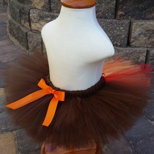 May include: A brown tutu skirt with an orange bow, displayed on a white mannequin torso with a wooden base. The tutu has layers of brown tulle with hints of red and orange.