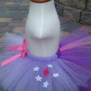 May include: A purple and white tutu skirt with a pink bow. The skirt is decorated with white and red star shapes. The tutu is displayed on a mannequin with a wooden head.