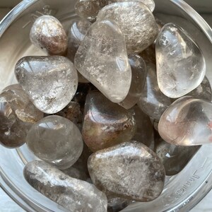 May include: A bowl filled with tumbled smoky quartz stones. The stones are a light grey color with a translucent appearance.