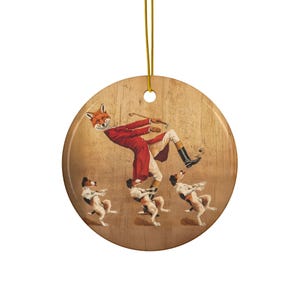 May include: A round ceramic ornament with a whimsical illustration of a fox-headed figure in a red coat falling, with three dogs. The ornament has a gold string for hanging and a wood-grain background.