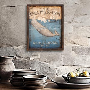 Puede incluir: Cartel enmarcado de madera con el texto "THE SPOUTER-INN NEW BEDFORD EST. 1851" que presenta una ballena y un barco sobre un fondo azul. El cartel está montado en una pared de piedra. Cerámica y platos decorativos en primer plano.