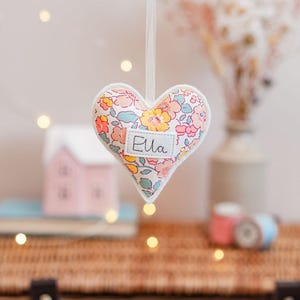 May include: A heart-shaped fabric ornament with a floral pattern in pink, yellow, and blue. The word "Ella" is printed on a white label. The ornament is hanging by a white ribbon, with a blurred background featuring a house and flowers.