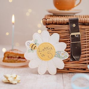 May include: A white daisy-shaped applique with the text "Lottie is 5" in a yellow center, and a small bee. A slice of cake with a lit candle, a wicker basket, and a teacup are in the background. The applique is likely for a birthday.