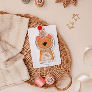 Può includere: Un biglietto di compleanno con un leone con un cappello da festa, con la scritta "Archie is 3". Il biglietto è su un cestino di vimini con rocchetti di filo, forbici e decorazioni a forma di stella. L'indirizzo del sito web è visibile.