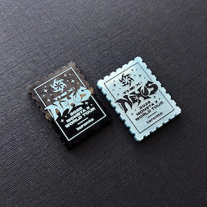 May include: Two rectangular postage stamp-shaped pins. One is black with silver text and graphics, the other is light blue with black text and graphics. Both feature the text THE X: NEXUS, 2026 MONSTA X WORLD TOUR, and toronto.
