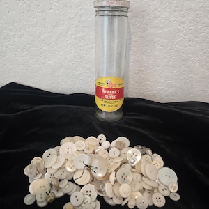 May include: A tall, cylindrical glass jar with a metal lid and a yellow and red label reading Albert's Olives, positioned behind a large, scattered pile of assorted white and off-white buttons of various sizes, shapes, and hole configurations.