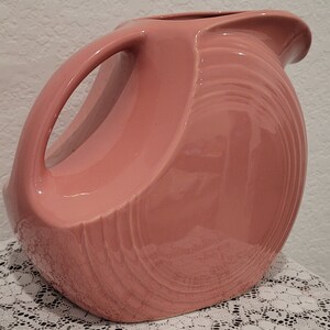 May include: A pink ceramic pitcher with a unique, curved design and a smooth, glossy finish. The pitcher has a wide mouth and a handle on the side.