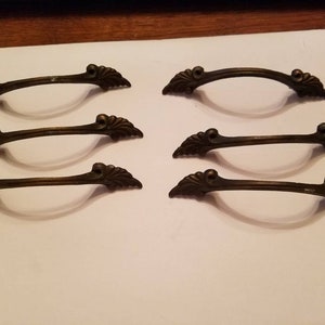 May include: Six antique bronze-toned drawer pulls with a decorative, ornate design. Each pull features a curved handle and decorative end pieces. The pulls are arranged on a white surface, showcasing their details.