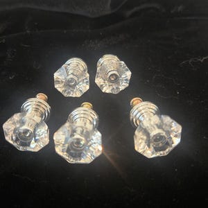 May include: Six clear, faceted glass cabinet knobs with silver-colored metal bases and gold-colored tops. The knobs are arranged on a dark surface, showcasing their reflective qualities and geometric design. These knobs are suitable for furniture restoration or new builds.