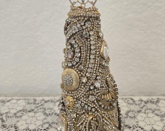 Puede incluir: Un adorno de árbol de Navidad de forma cónica dorada cubierto de diamantes de imitación y otros adornos decorativos. La parte superior del árbol está adornada con una estrella dorada de filigrana.