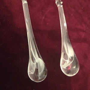 May include: Two clear glass chandelier crystals with a teardrop shape. Each crystal has a white, etched floral design inside. The crystals hang from gold-colored metal fixtures against a dark red background.