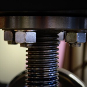 May include: Close-up of a metal screw mechanism with a large, silver-colored nut on top. The screw is made of multiple stacked metal rings.