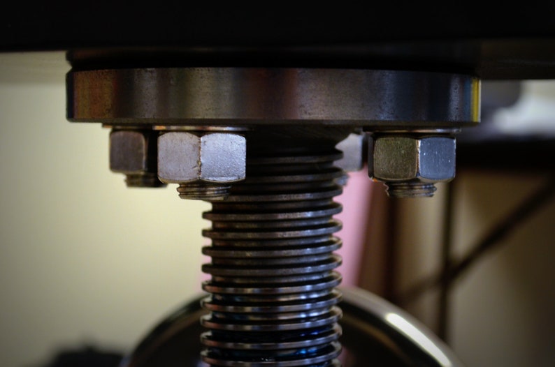 May include: Close-up of a metal screw mechanism with a large, silver-colored nut on top. The screw is made of multiple stacked metal rings.