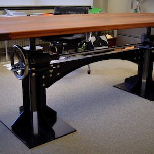 May include: A wooden desk with a black metal frame and adjustable height. The desk has a crank on the side to adjust the height. The desk is in a room with a carpeted floor.