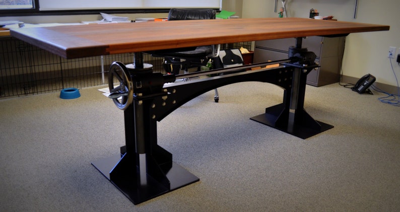May include: A wooden desk with a black metal frame and adjustable height. The desk has a crank on the side to adjust the height. The desk is in a room with a carpeted floor.