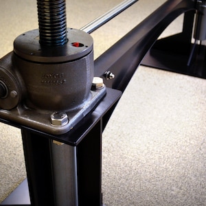 May include: Close-up of a black metal table leg with a silver adjustable height mechanism. The mechanism has a black base with a silver screw and a silver rod. The base is labeled "JOYCE TECHNOLOGY".