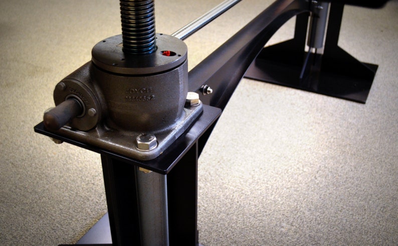 May include: Close-up of a black metal table leg with a silver adjustable height mechanism. The mechanism has a black base with a silver screw and a silver rod. The base is labeled "JOYCE TECHNOLOGY".