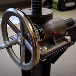 May include: A silver metal hand crank with a black base and a silver metal rod extending from the side. The crank has a large silver wheel with a black handle. The base has a black metal plate with a silver metal label that reads "JOYCE SERIAL NO. 1124466".