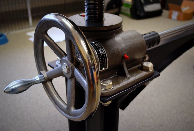 May include: A silver metal hand crank with a black base and a silver metal rod extending from the side. The crank has a large silver wheel with a black handle. The base has a black metal plate with a silver metal label that reads "JOYCE SERIAL NO. 1124466".