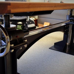 May include: A black metal desk base with a wooden top. The base has a crank handle and a metal rod that connects the two sides. The desk base is on a carpeted floor.