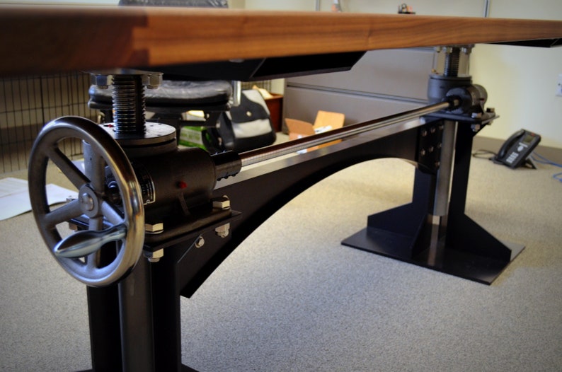 May include: A black metal desk base with a wooden top. The base has a crank handle and a metal rod that connects the two sides. The desk base is on a carpeted floor.