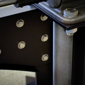 May include: Close-up of a black metal bracket with multiple silver bolts attaching it to a silver metal pole. The bracket has a rounded edge and is attached to a larger metal structure.
