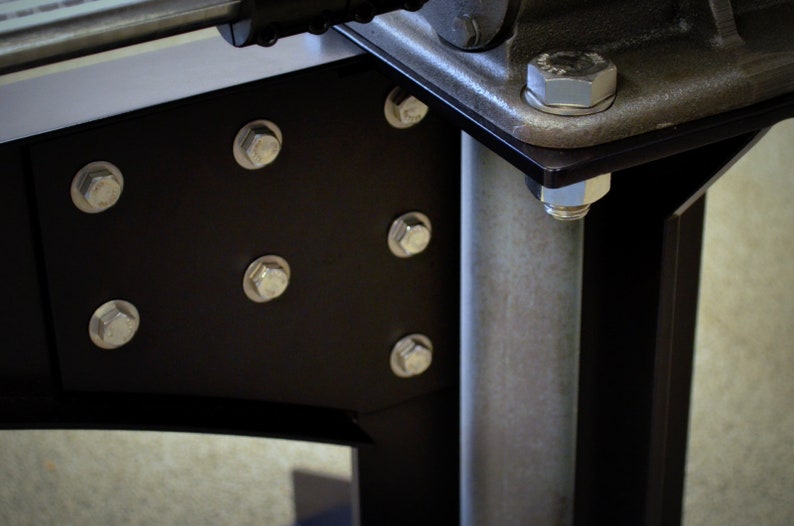 May include: Close-up of a black metal bracket with multiple silver bolts attaching it to a silver metal pole. The bracket has a rounded edge and is attached to a larger metal structure.