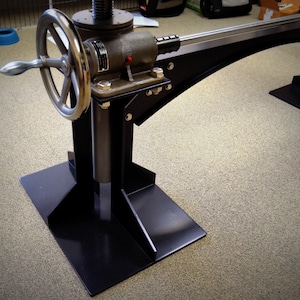May include: A black metal stand with a large wheel and a lever. The stand has a base with three legs and a vertical post with a threaded rod. The stand is used to adjust the height of a table or other object.