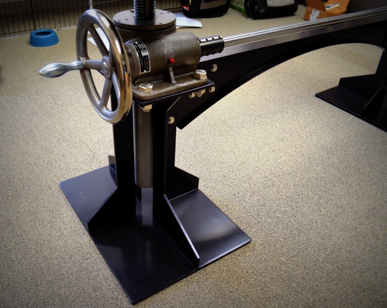 May include: A black metal stand with a large wheel and a lever. The stand has a base with three legs and a vertical post with a threaded rod. The stand is used to adjust the height of a table or other object.