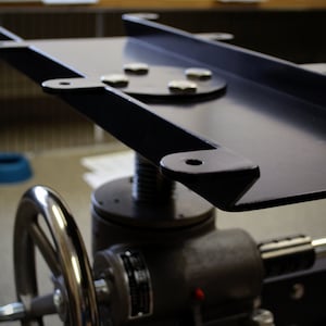 May include: A black metal platform with a round, silver-colored base and a hand crank. The platform is attached to a black metal frame with a screw mechanism.