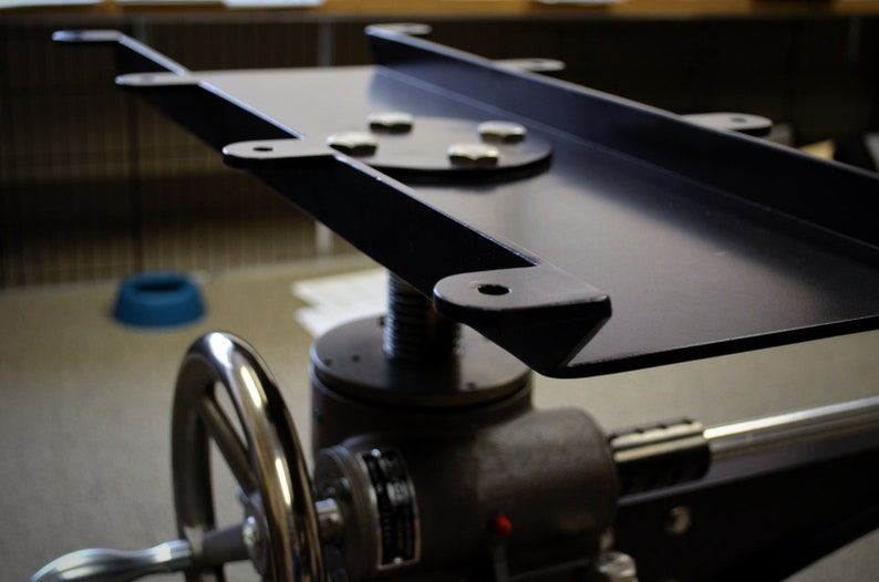 May include: A black metal platform with a round, silver-colored base and a hand crank. The platform is attached to a black metal frame with a screw mechanism.