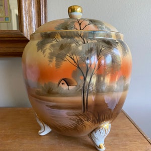 May include: A ceramic cookie jar with a lid, featuring a hand-painted landscape scene with a house, trees, and a river. The jar has three feet and is decorated with gold accents.