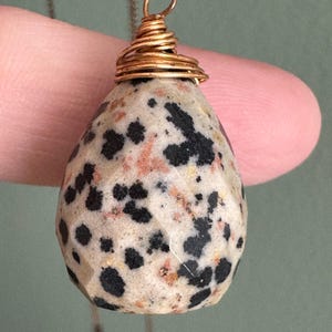 Puede incluir: Un colgante en forma de l&aacute;grima con un patr&oacute;n moteado de manchas negras sobre una piedra de color crema. La piedra est&aacute; envuelta con alambre de color dorado y suspendida de una delicada cadena. El colgante est&aacute; sujeto por un dedo.