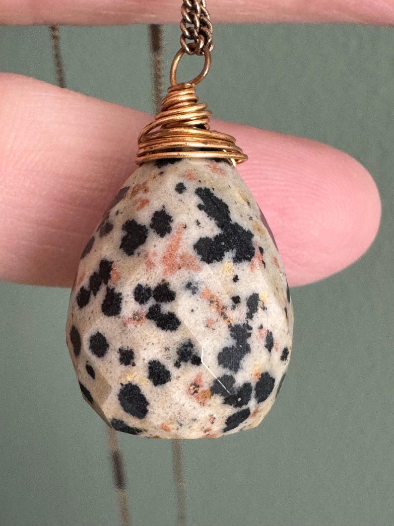 Puede incluir: Un colgante en forma de l&aacute;grima con un patr&oacute;n moteado de manchas negras sobre una piedra de color crema. La piedra est&aacute; envuelta con alambre de color dorado y suspendida de una delicada cadena. El colgante est&aacute; sujeto por un dedo.