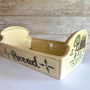 May include: A ceramic bread basket with a yellow glaze and a green and brown design. The basket has a raised edge and the word "Bread" is written on the front.