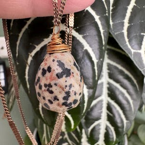 Puede incluir: Un collar con colgante de piedra moteada y cadena dorada. La piedra, en forma de l&aacute;grima, presenta manchas negras y rojizas sobre un fondo beige. La piedra est&aacute; envuelta con alambre dorado.