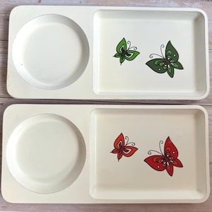 May include: Two off-white serving trays. Each tray has a circular indentation and a rectangular section decorated with butterfly illustrations. One tray has green butterflies, the other has red butterflies.