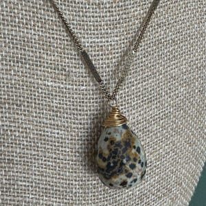 Puede incluir: Un collar con colgante en forma de l&aacute;grima. El colgante es una piedra moteada, envuelta con alambre dorado. La cadena es de color dorado. El collar se exhibe sobre una superficie texturizada de color neutro.